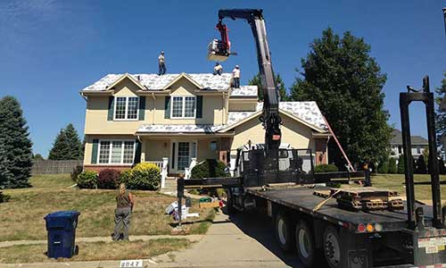 Excalibur Roofing crew members on a roof installing new asphalt shingles, a large truck with a crane is delivering shingle packs to the team on the roof
